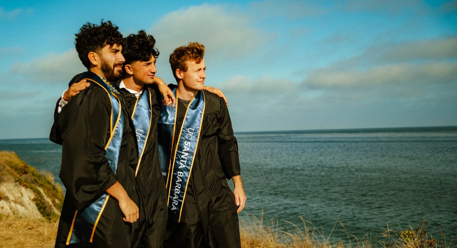 Daniel with friends in UC Santa Barbara graduation gowns and blue sashes, arms around each other, on a bluff above the Pacific.